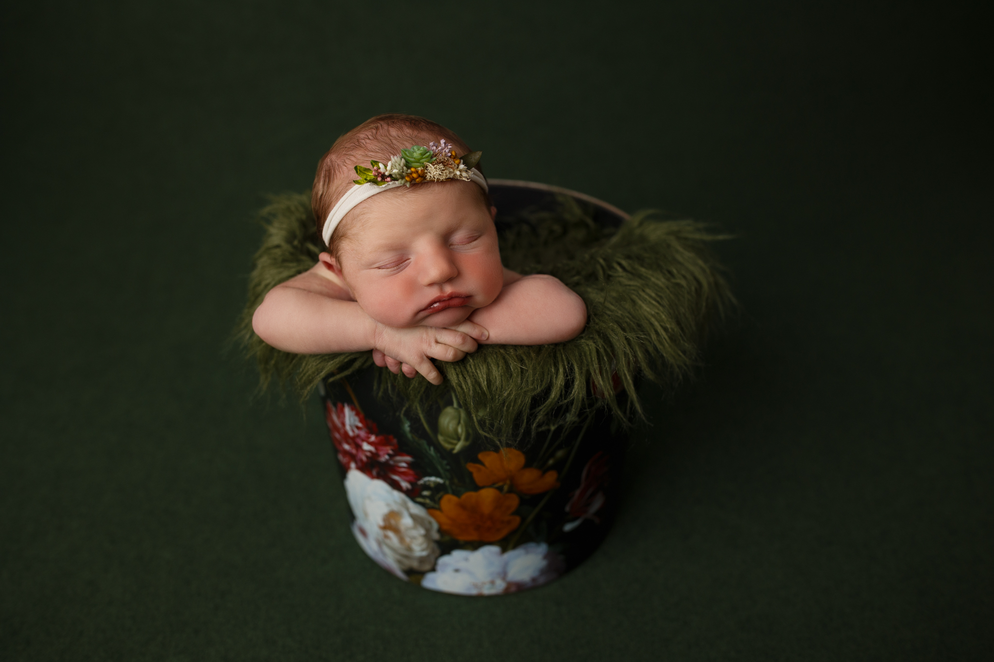 A sleeping baby girl in a floral bucket with a dark green background.