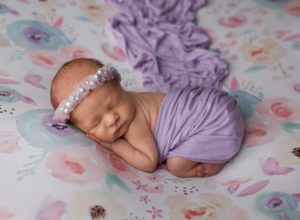 A newborn baby lies on her tummy on a floral backdrop with a beaded purple headband.